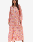 A woman modeling a long-sleeve, floor-length kaftan dress with a peach and pink paisley pattern, set against a plain light gray background.