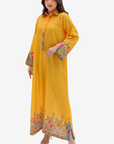 A woman modeling a long yellow kaftan dress with colorful floral embroidery on the cuffs and hem, set against a plain light gray background.