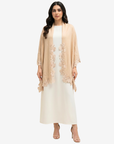 A woman modeling a light beige open-front cardigan with floral lace trim over a white maxi dress, standing against a plain light gray studio background.