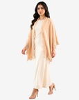 A woman modeling a light peach-beige open-front shawl or wrap with floral lace trim over a cream maxi dress, against a plain light gray studio background.