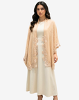 A woman modeling a light peach-beige open-front shawl or wrap with floral lace trim over a cream midi dress, set against a plain light gray studio background.