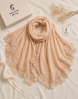 A peach-colored shawl scarf with intricate floral lace trim, elegantly draped on a white fabric background alongside a branded white box, a ribbon, and dried flowers. The brand 'GAMZA COUTURE' is visible on the packaging and ribbon.