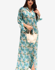 A woman modeling a long-sleeved, floor-length floral maxi dress with a high slit, holding a cream-colored handbag against a solid light gray background.