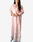 A woman modeling a long light pink kaftan dress with a floral print concentrated along the hem and small embroidered details near the neckline and cuffs, set against a plain light gray background.