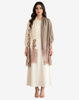 A woman modeling a taupe shawl with rose gold floral lace borders over a cream-colored dress, set against a plain light gray background.