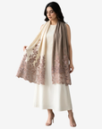 A woman models a long, lightweight shawl draped over a cream-colored dress. The shawl features a gradient transition from cream at the top to a dusty rose-beige at the bottom, with detailed floral embroidery concentrated along the lower edge.