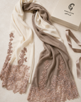 A draped scarf in cream and taupe with rose gold floral lace embroidery, displayed next to a beige gift box on a neutral beige background.