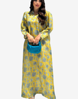 A woman modeling a long yellow kaftan dress with a gray palm tree pattern, holding a small blue handbag against a light gray background.