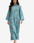 A woman modeling a long-sleeved, collared maxi dress in turquoise blue with a brown and beige floral pattern, set against a plain light gray background.