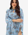 A woman modeling a light blue long-sleeve tunic dress with a gray palm tree and floral pattern, featuring a keyhole neckline and tassel tie details.