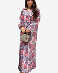 A woman modeling a long-sleeved, collared maxi dress with a pink, purple, and white abstract pattern, holding a small striped handbag against a light gray background.