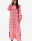 A woman modeling a long pink and white printed kaftan dress with geometric patterns, 3/4 sleeves, and side tie details, set against a plain light gray background.