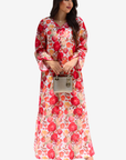 A woman modeling a long-sleeved maxi dress with a vibrant red, pink, and orange floral print on a white base, holding a small metallic handbag against a light gray background.