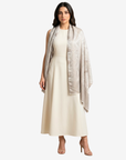 A woman modeling a cream midi dress with a light beige patterned shawl draped over her shoulders, set against a plain light gray studio background.