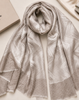 A light beige and silver patterned scarf draped elegantly on a cream surface, styled with a beige brand box, a clutch, dried flowers, and a small branded card. The box and card display the brand name 'GAMREA COUTURE'.