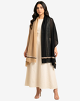 A woman modeling a black sheer shawl with a beige and black zigzag border over a cream-colored dress, set against a plain light gray background.