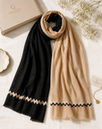 Two lightweight scarves from Gharza London are displayed in a flat-lay arrangement on a textured white fabric background. One scarf is solid black with a beige zigzag border, and the other is solid beige with a black zigzag border.