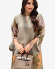 A woman modeling a loose, short-sleeved tunic dress in a muted sage green and beige floral print, holding a structured taupe handbag with silver hardware against a plain light gray background.