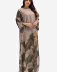 A woman modeling a long, loose-fitting maxi dress with a muted taupe base and an abstract landscape-style print in olive green, brown, and tan, shown against a plain light gray studio background.