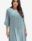 A woman modeling a light blue-green kaftan dress with intricate geometric embroidery in matching and dark purple tones on the chest and sleeves, set against a plain light gray background.
