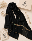A black sheer scarf with gold trim and green-and-gold embroidered geometric accents, styled in a flat lay with a branded cream box, drawstring pouch, tassel tag, and dried flowers on a light beige background.