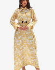 A woman modeling a long-sleeved yellow and white tropical print maxi dress with a matching belt featuring two black buttons, set against a plain light gray background.