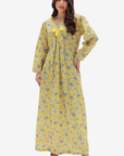 A woman modeling a long yellow maxi dress with a blue floral pattern and a matching yellow bow at the neckline, set against a plain light gray background.