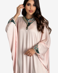 A woman modeling a light pink kaftan dress with embroidered floral details on the collar and cuffs, set against a plain light gray background.