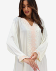 A woman wearing a long-sleeved white kaftan-style tunic with delicate pale pink floral lace embroidery along the V-neckline and cuffs, set against a plain light gray studio background.
