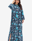 A woman modeling a long navy blue kaftan dress with a vibrant turquoise, white, and light pink floral and vine pattern, set against a plain light gray background.