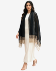 A woman modeling a black sheer shawl with beige lace trim over a cream-colored dress, set against a plain light gray background.