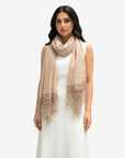 A woman modeling a light beige scarf with delicate lace trim at the ends, worn over a white sleeveless dress against a plain light gray background.