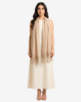 A woman modeling a long, lightweight beige scarf with delicate lace trim at the bottom, draped over a cream-colored sleeveless dress against a light gray background.