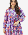 A woman wearing a purple long-sleeve kaftan dress with a colorful floral print in pink, orange, and white, set against a plain light gray background.