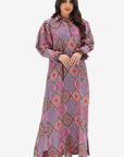 A woman modeling a long-sleeved, collared maxi dress with a vibrant pink, purple, and orange paisley pattern against a plain light gray background.