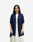 A woman modeling a navy blue open-front shawl or lightweight jacket with decorative geometric embroidery down the front, worn over a white dress against a light gray studio background.