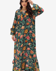 A woman modeling a long-sleeved maxi dress with a vibrant floral print on a dark green background. The dress features orange, pink, and yellow flowers with green leaves, and has a split hem with yellow piping.
