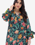 A woman wearing a long-sleeve floral print tunic dress with green leaves, pink and orange flowers, and yellow trim on the cuffs, set against a plain light gray background.