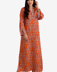 A woman modeling a long orange kaftan dress with a gray geometric pattern and white piping details, set against a plain light gray background.