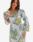 A woman modeling a long-sleeve midi dress with a white base and a vibrant lemon, blue bird, and floral print, set against a plain light gray background.