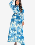 A woman modeling a long-sleeve, collared maxi dress with a blue and white palm tree print, featuring a matching belt with double gold-tone buckles, set against a plain light gray background.