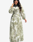 A woman modeling a long-sleeved maxi dress with a green botanical and scroll pattern on a white background, set against a plain light gray studio backdrop.