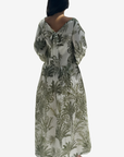 A woman viewed from the back wearing a long-sleeve maxi dress with a tie-neck detail and an all-over botanical print in olive green on a light beige background.