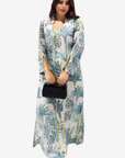 A woman modeling a long-sleeved maxi dress with a blue and green botanical print on a white base, featuring yellow piping details, holding a small black handbag against a light gray background.