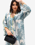 A woman modeling a long-sleeve white kaftan-style tunic with a blue tropical palm tree print and yellow piping trim, holding a black handbag against a light gray studio background.