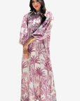 A woman modeling a long-sleeved maxi dress with a pink and white palm tree pattern, set against a plain light gray background.