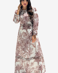 A woman modeling a long-sleeved maxi dress with a brown palm tree and floral pattern on a cream background, set against a plain light gray studio background.