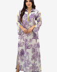 A woman modeling a long-sleeved maxi dress with a purple palm tree pattern on a white background, featuring yellow piping details along the collar, cuffs, and side slits.