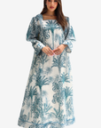 A woman modeling a long-sleeved maxi dress with a blue and white tropical palm tree print, set against a plain light gray background.