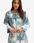 A woman wearing a long-sleeve dress with a blue palm tree toile pattern on a white background, set against a plain light gray studio backdrop.